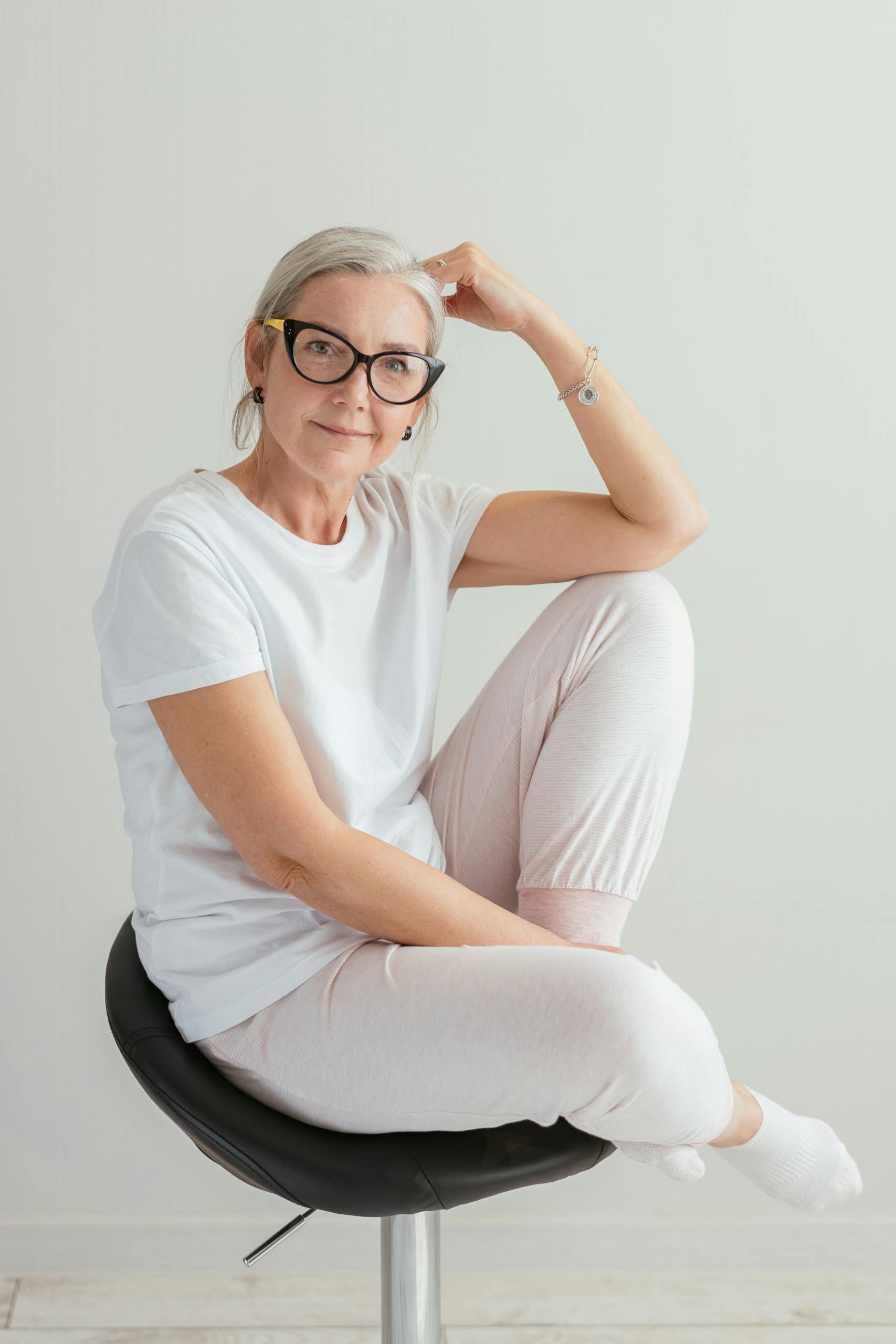 Precision Metabolic Care for Women Hawaii. A senior woman with eyeglasses smiling in a relaxed pose, wearing casual white clothes, seated indoors.