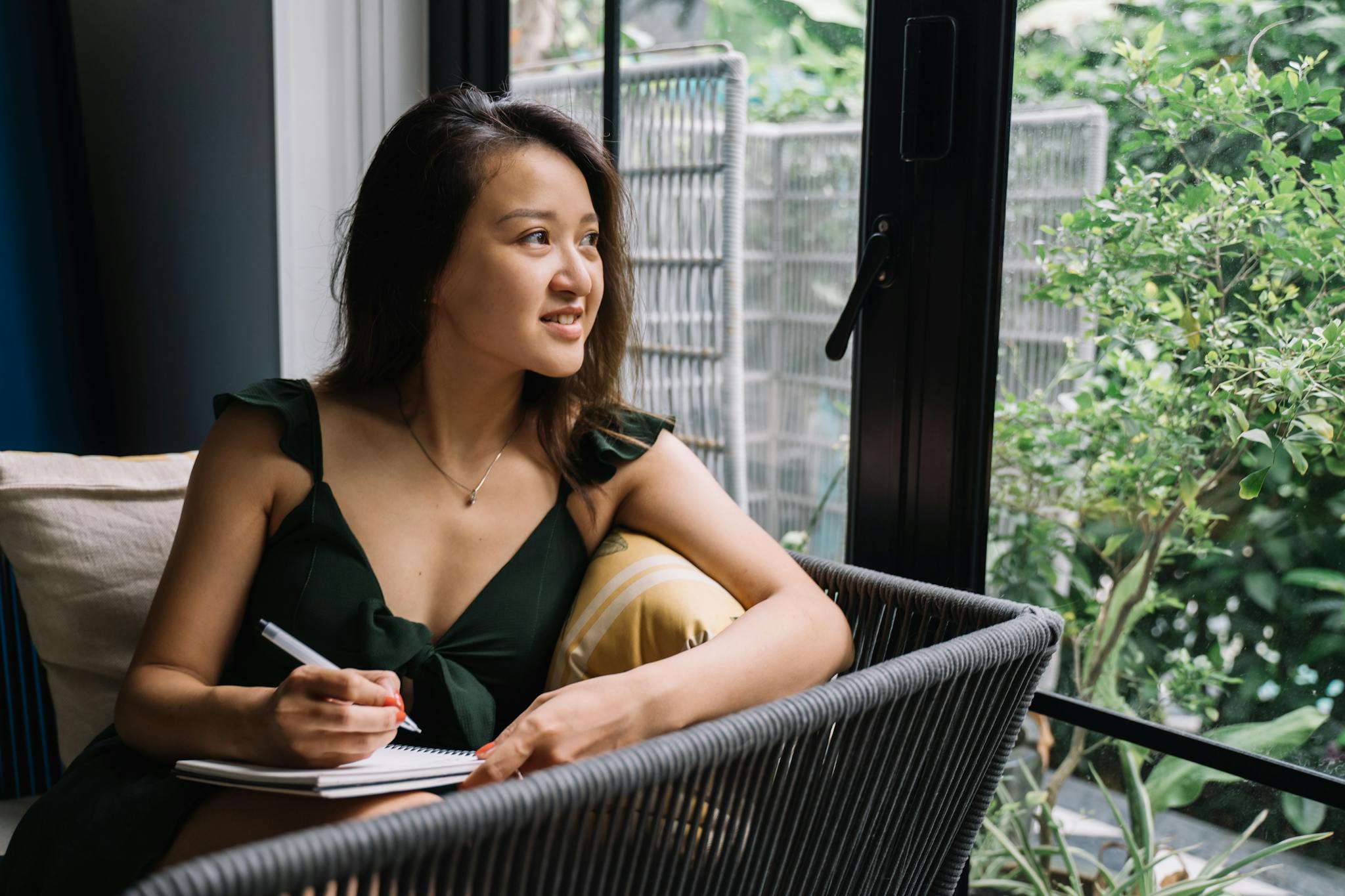 Asian woman in green dress writing in a notepad by the window, smiling and relaxed.