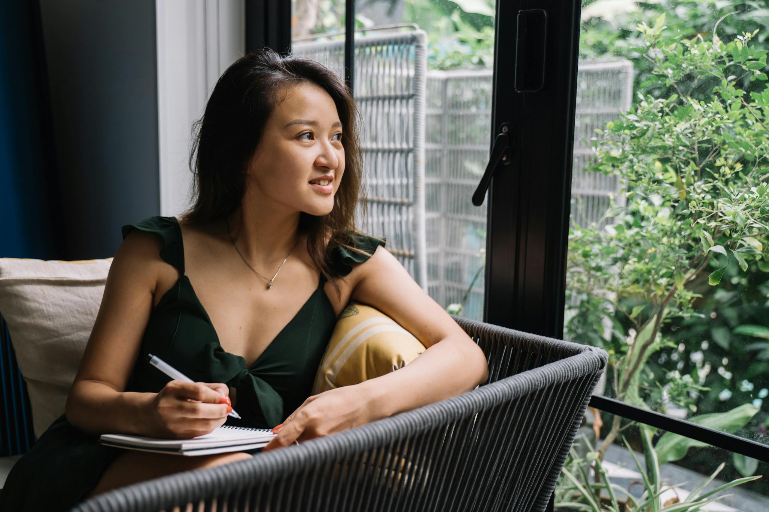 Asian woman in green dress writing in a notepad by the window, smiling and relaxed.