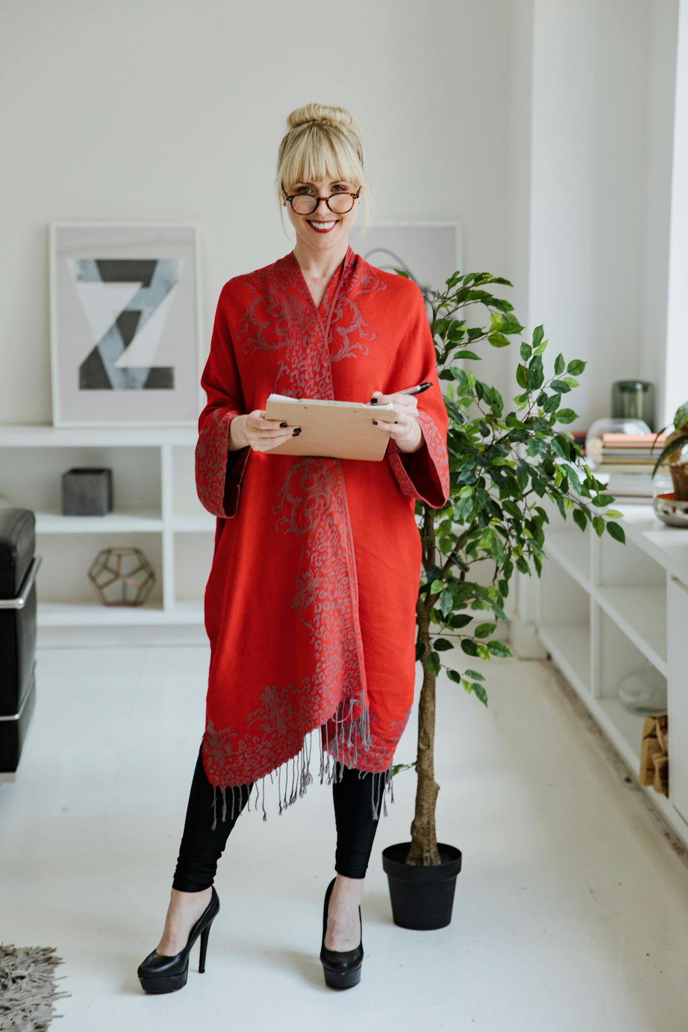 Blonde woman wearing glasses and red robe smiles while holding a tablet indoors.