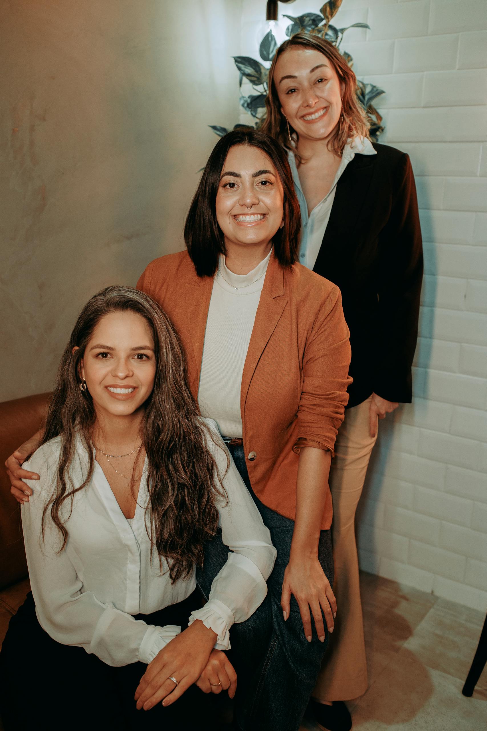 Three women in professional attire smiling indoors, embodying elegance and confidence in a modern setting.
