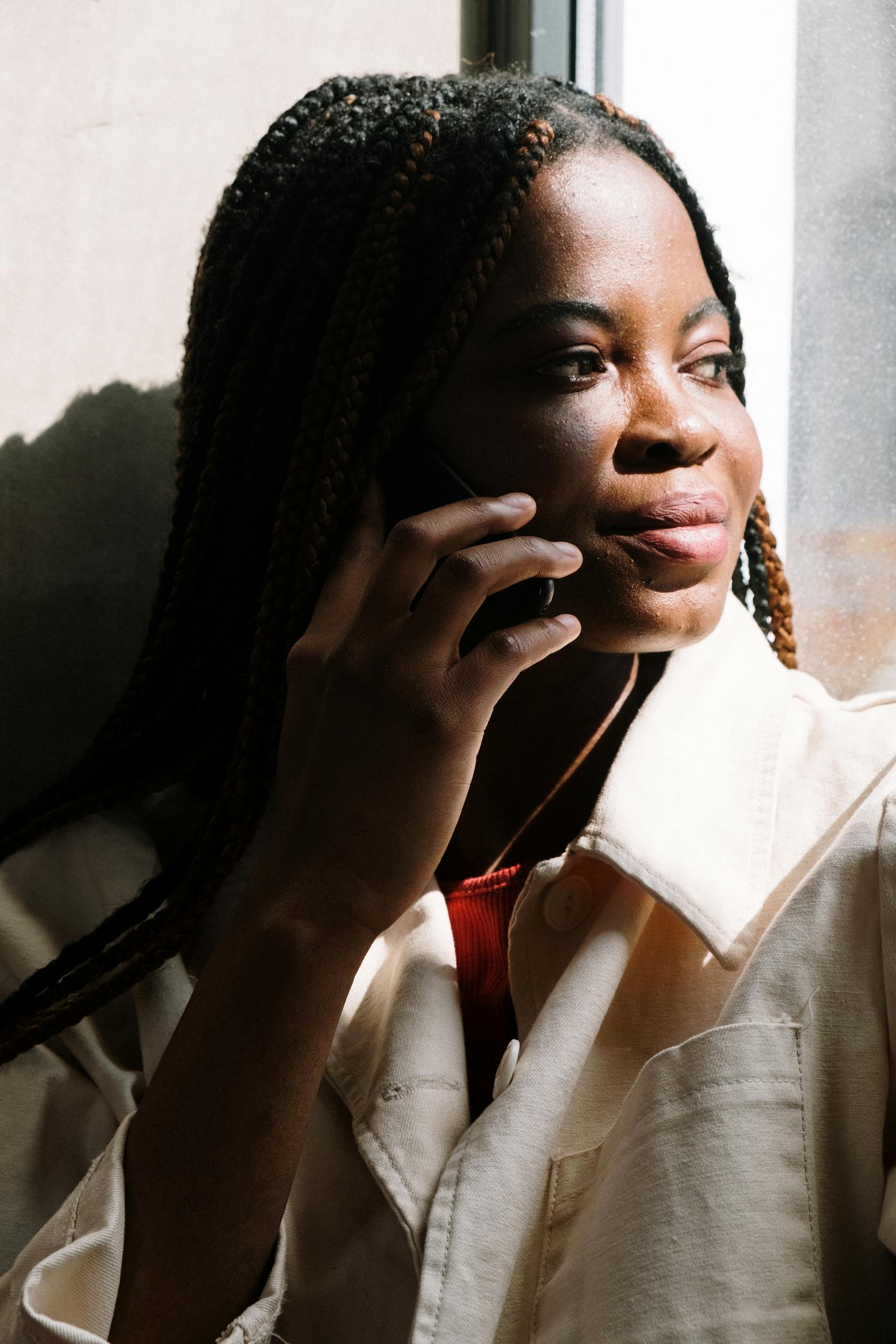 A woman with braided hair talking on a cellphone by a sunny window, showcasing modern communication.